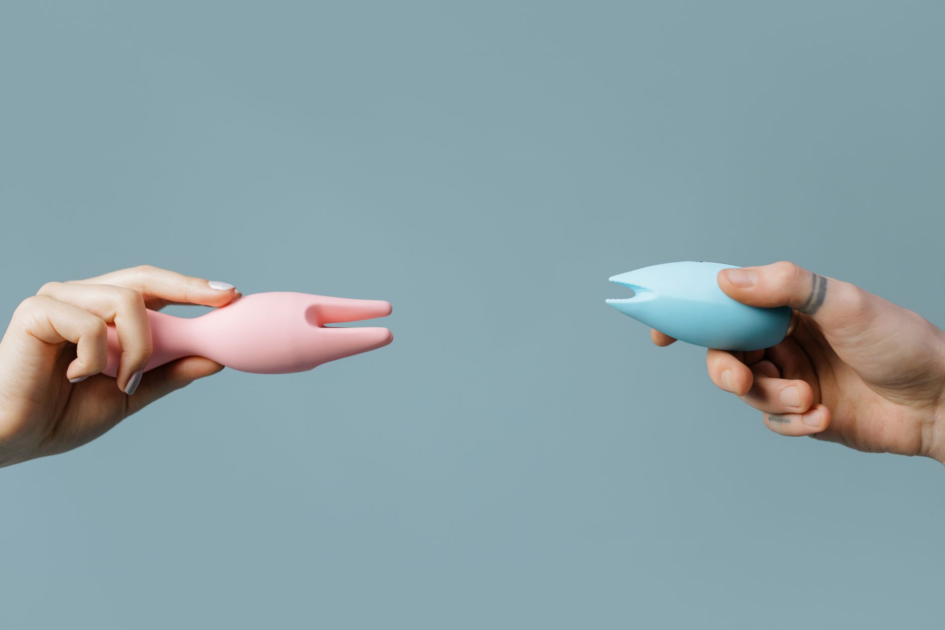 Manos sosteniendo vibradores de silicona rosa y azul contra fondo pastel minimalista